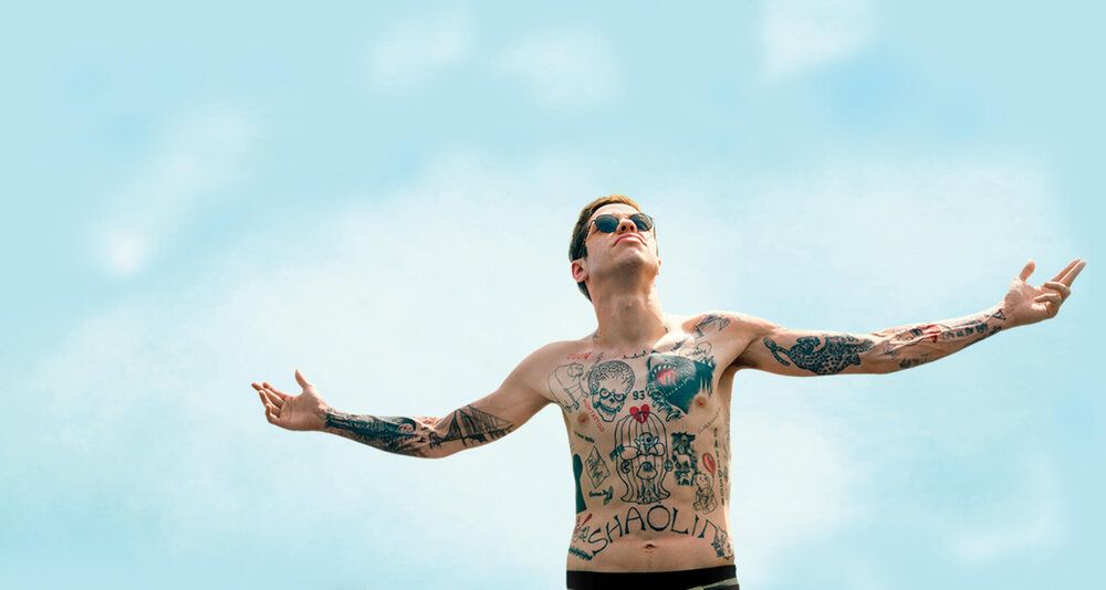 Heavily tattooed and bare-chested man stands with arms outstretched with blue sky behind him