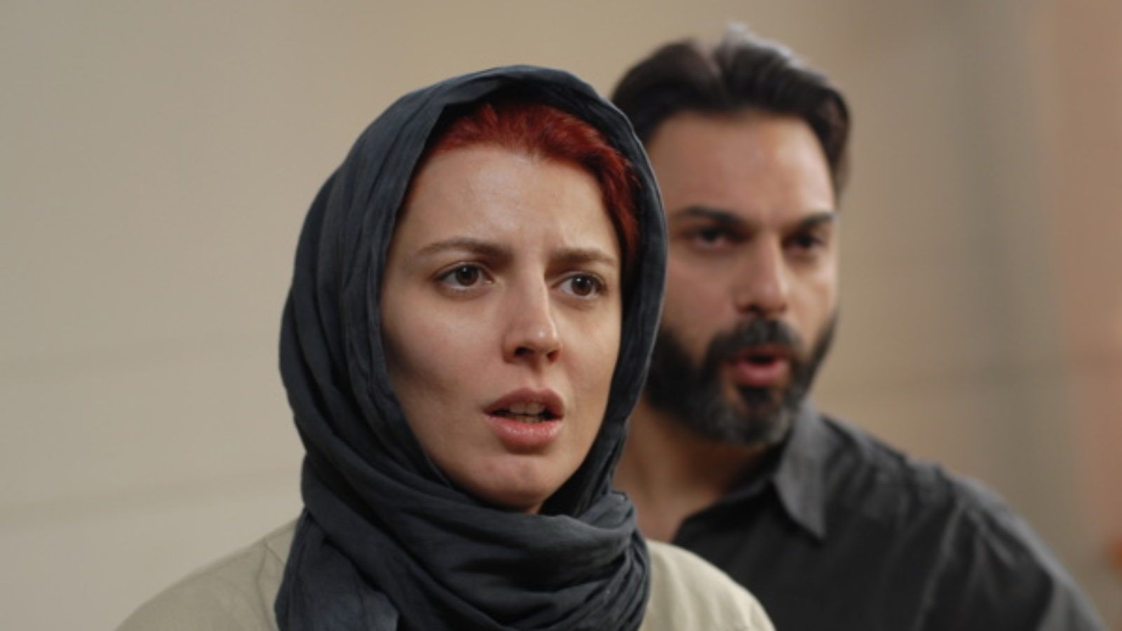 Woman in hijab stands in front of man; she looks concerned.
