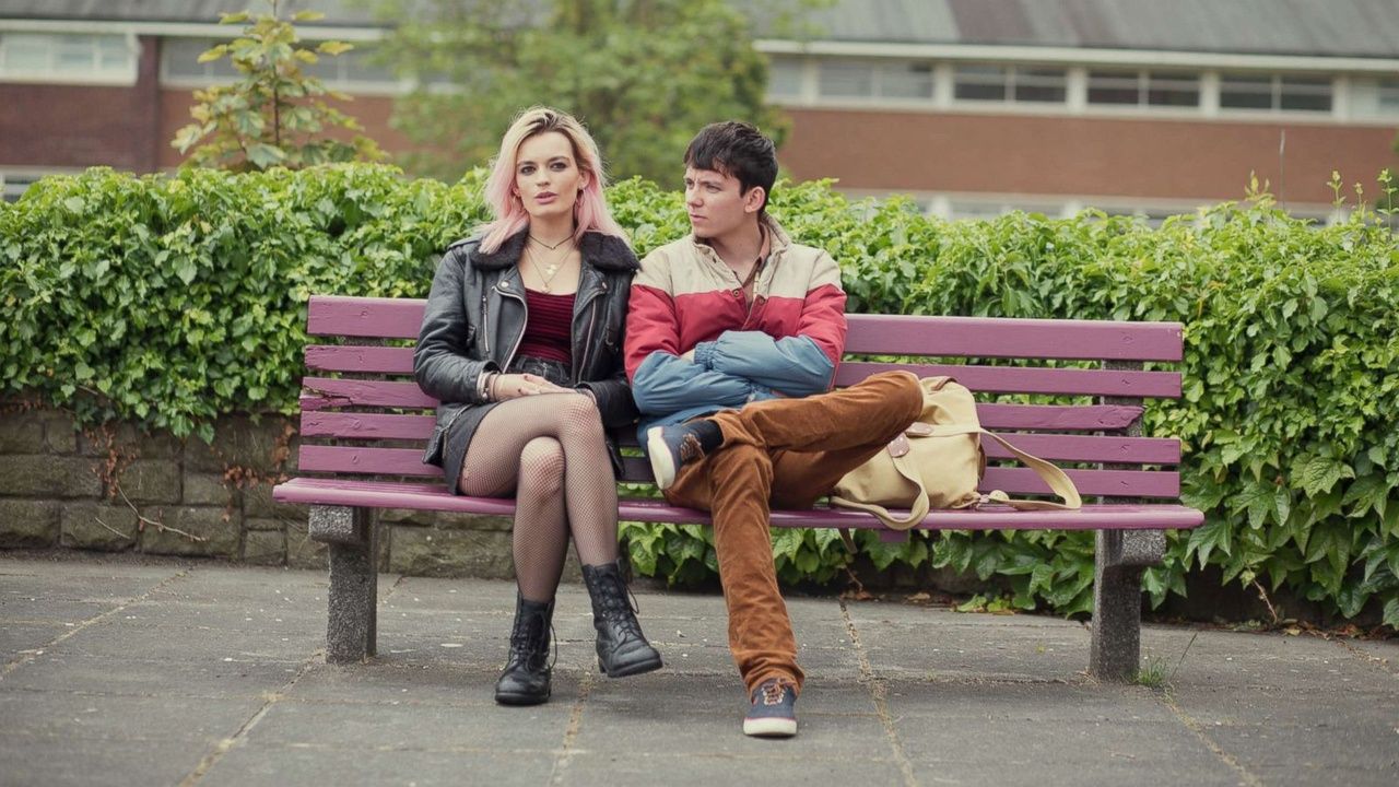The two teenage characters of Sex Education sit on a pink bench