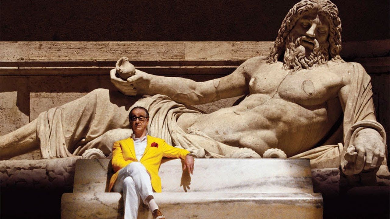 Man in yellow suit jacket sits in front of Roman monument. 