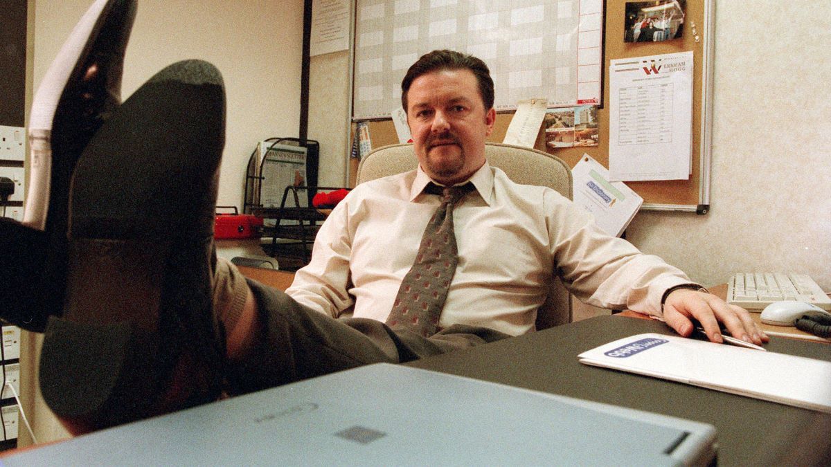 Ricky Gervais propping his feet up on an office desk.