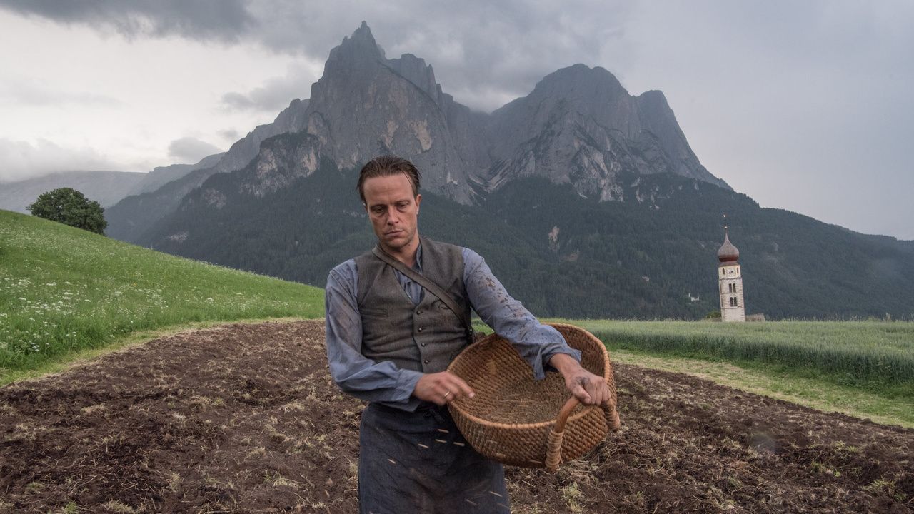 A farmer dejectedly carrying an empty basket along a dirt road, a large mountain behind him