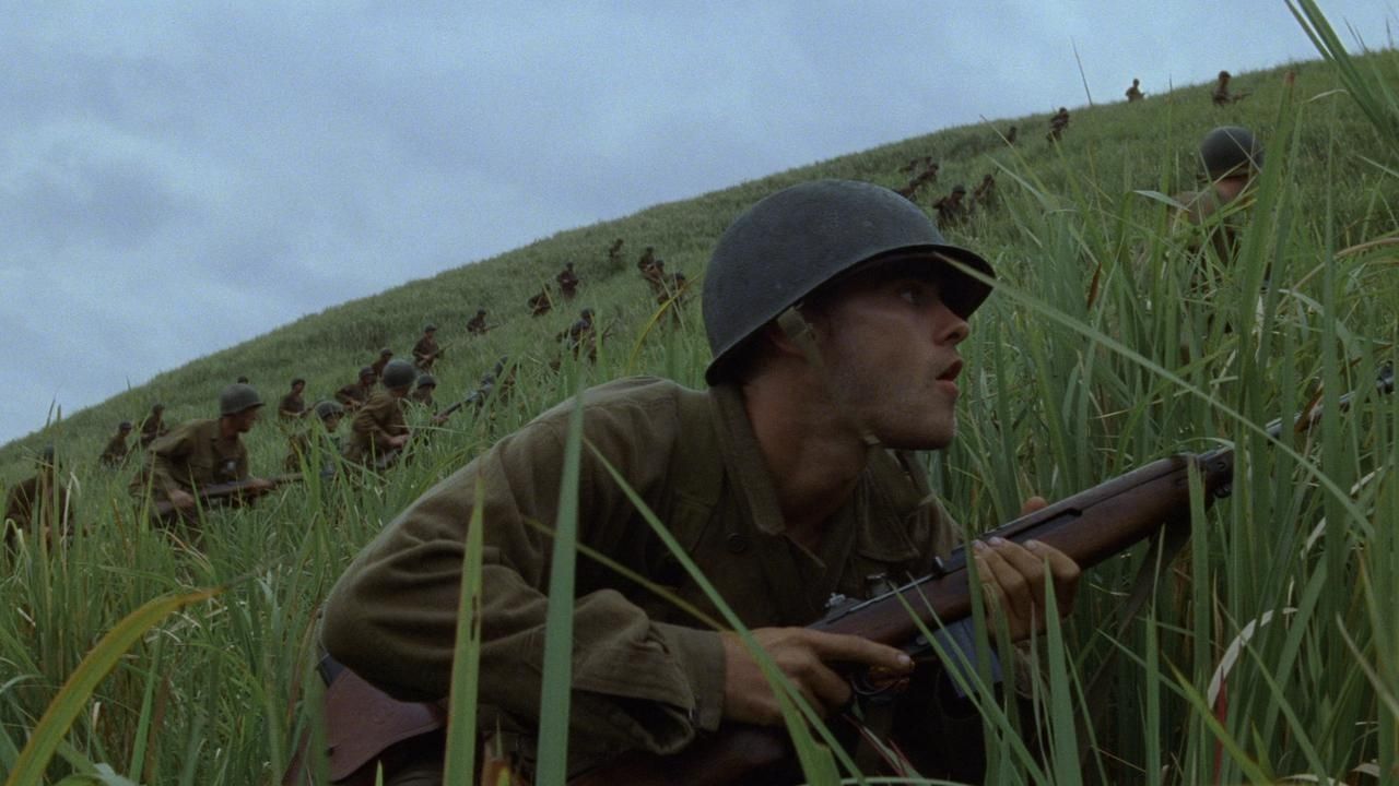 A soldier crouching low in the grass as dozens of other soldiers dot the hill behind him