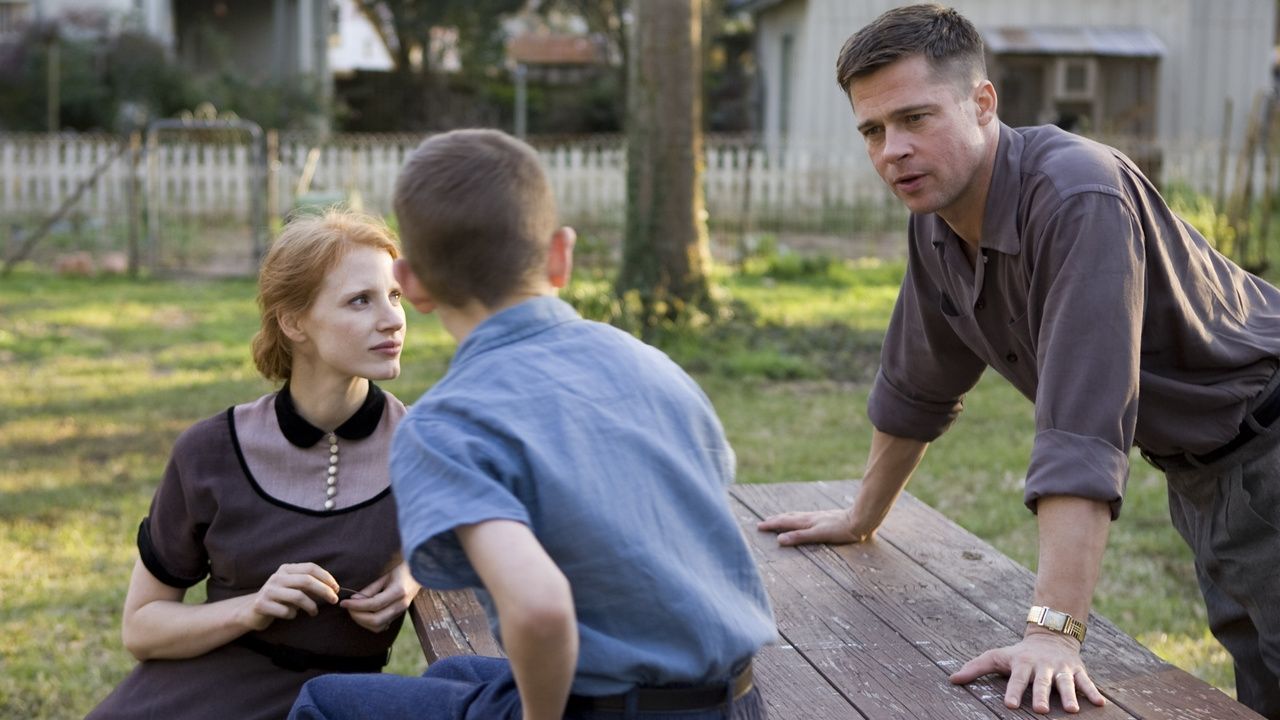 Brad Pitt and Jessica Chastain on either side of a picnic table as a child sits on the table, looking at them and facing away from the camera.