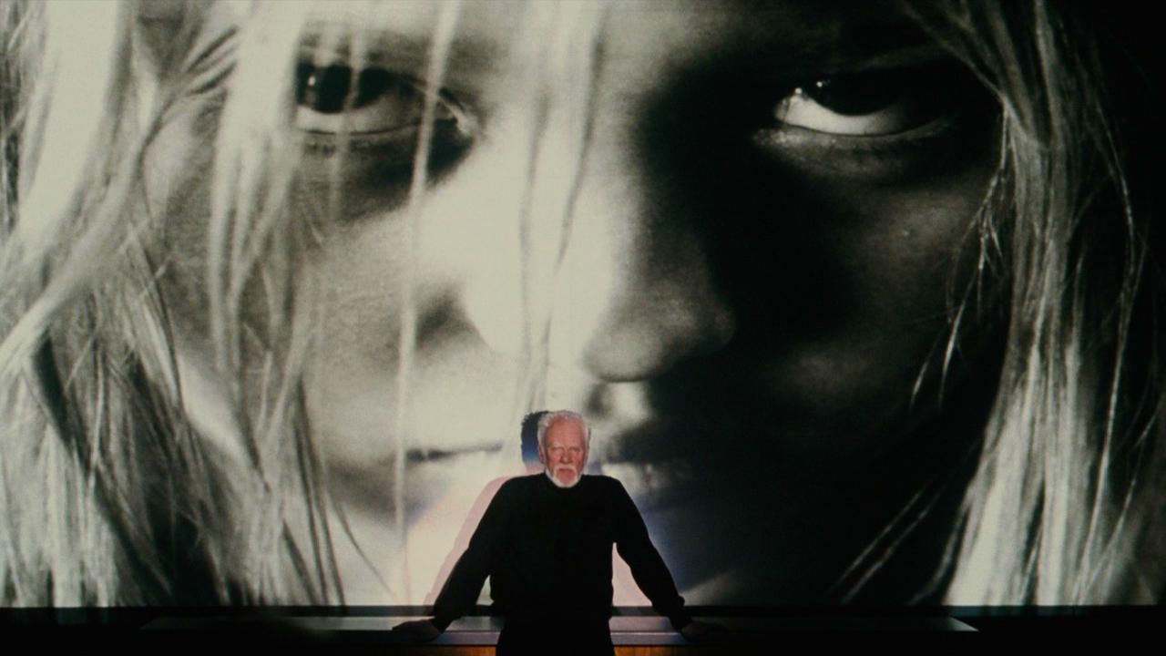 Malcolm McDowell stands in front of a big screen blow up of a disturbed child