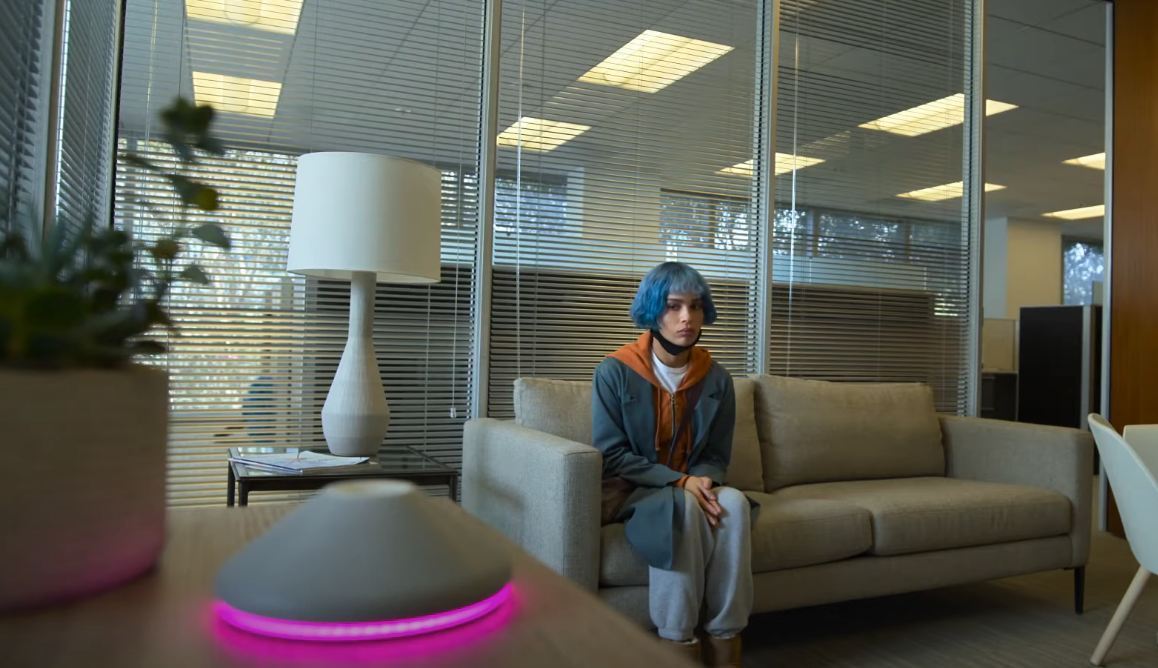 Woman with blue hair and orange hoodie sits on couch.