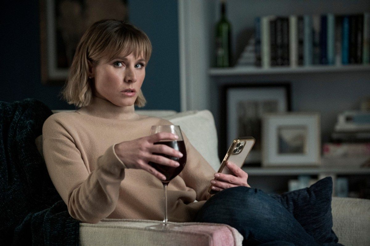 Kristen Bell relaxing with a wine glass in one hand and a phone in the other.