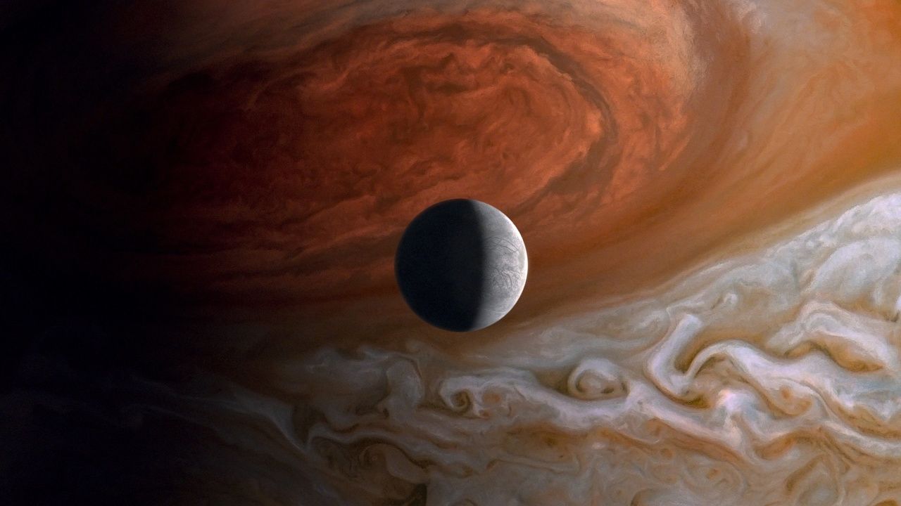 a small moon is partially in darkness as Jupiters great red spot looms in the background.