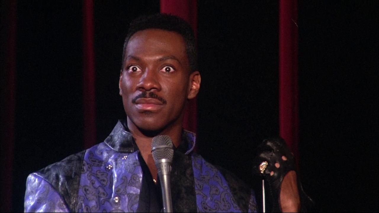 Eddie Murphy deadpans to the camera with microphone in hand and purple outfit