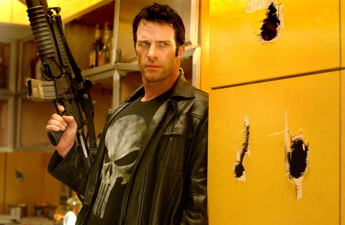 Thomas Jane as the Punisher, a large gun in his hand as he hides around a corner, the wall next to him filled with several large bullet holes.