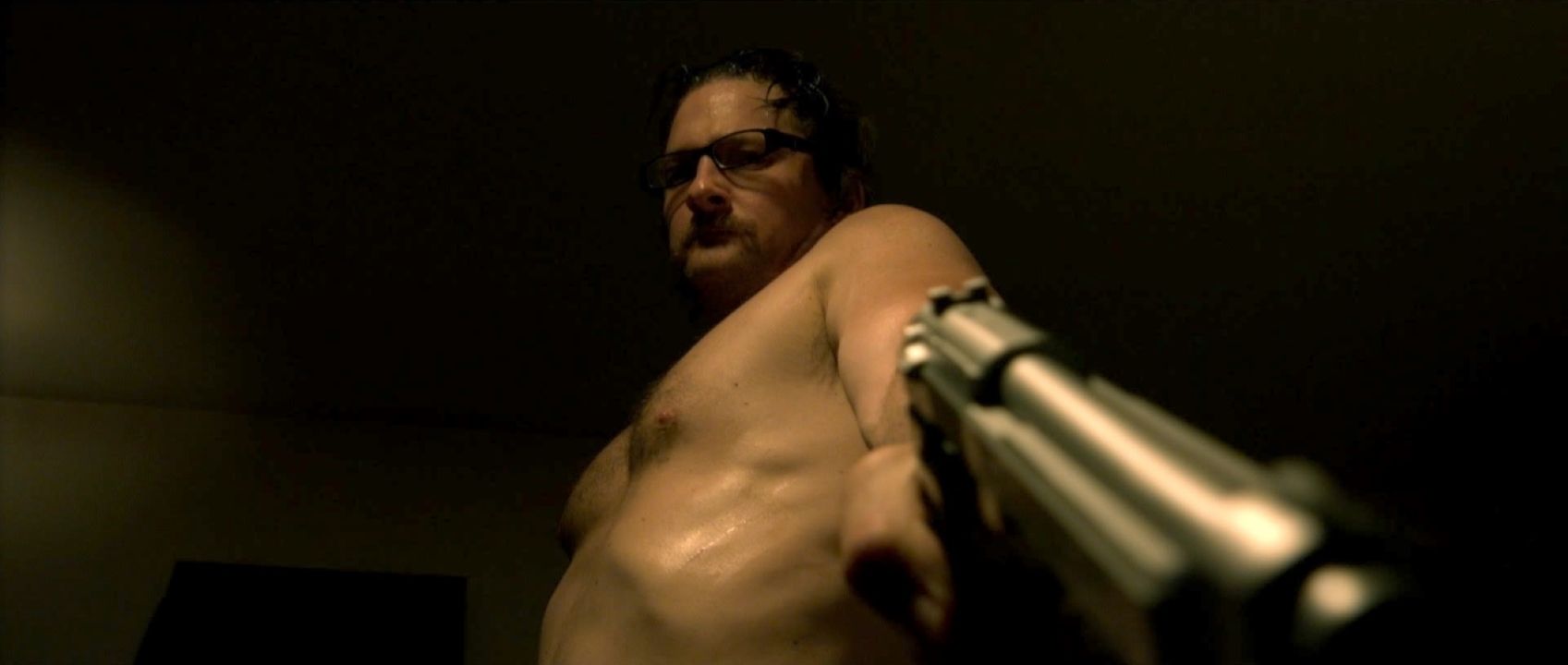 A shirtless Ray Belfi points a gun downward