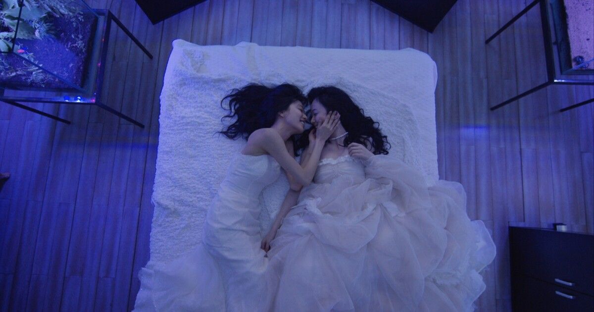 Two women sit in bed together while in wedding dresses.