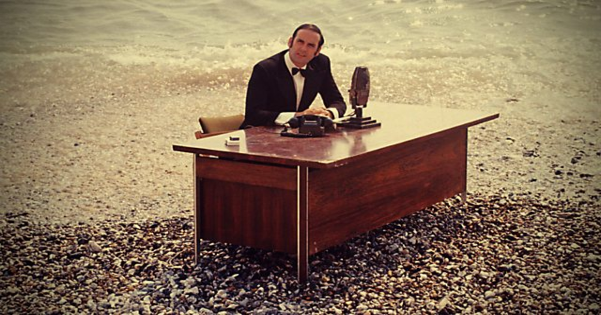 John Cleese at a desk on the beach in And Now for Something Completely Different