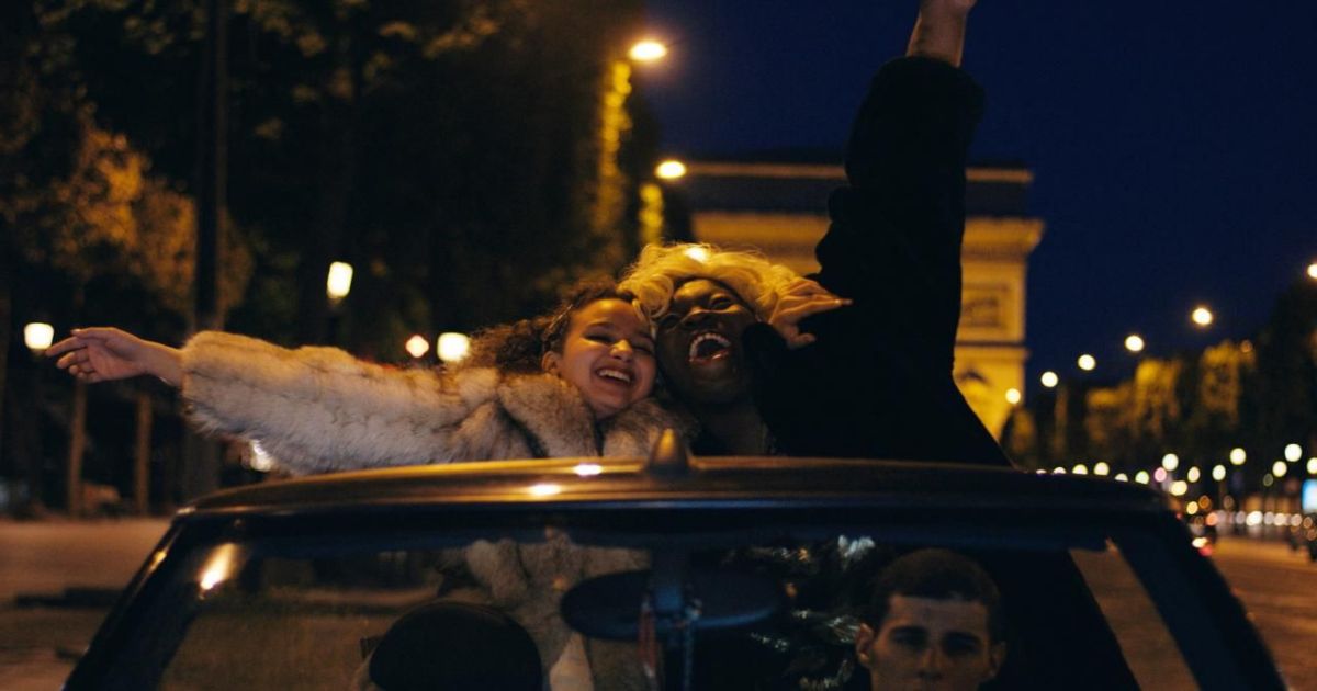Two girls holding their arms up in a convertible driving down the champs elysses in Divines