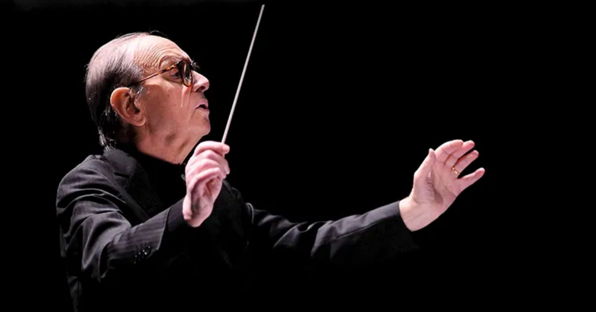 Ennio Morricone wearing black in a black background conducting an offscreen orchestra