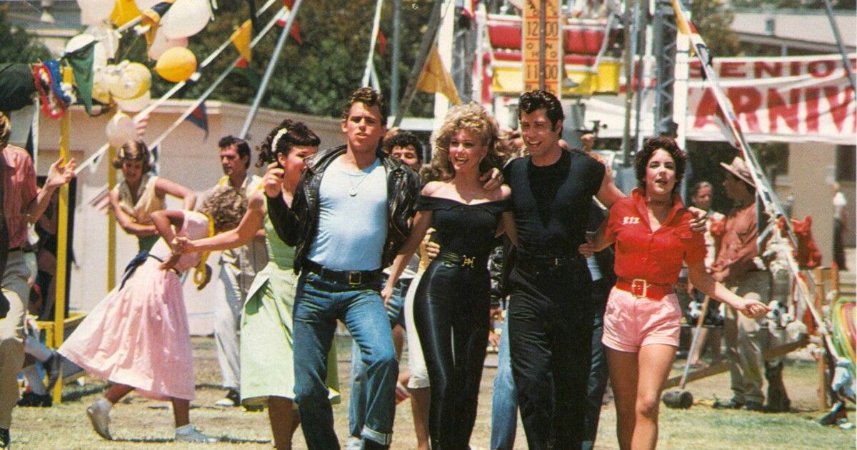 The cast of Grease linking arms and walking together, while laughing.