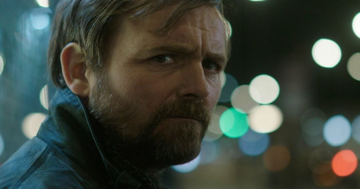 Neil Maskell looks sadly at the camera with blurred city lights behind him