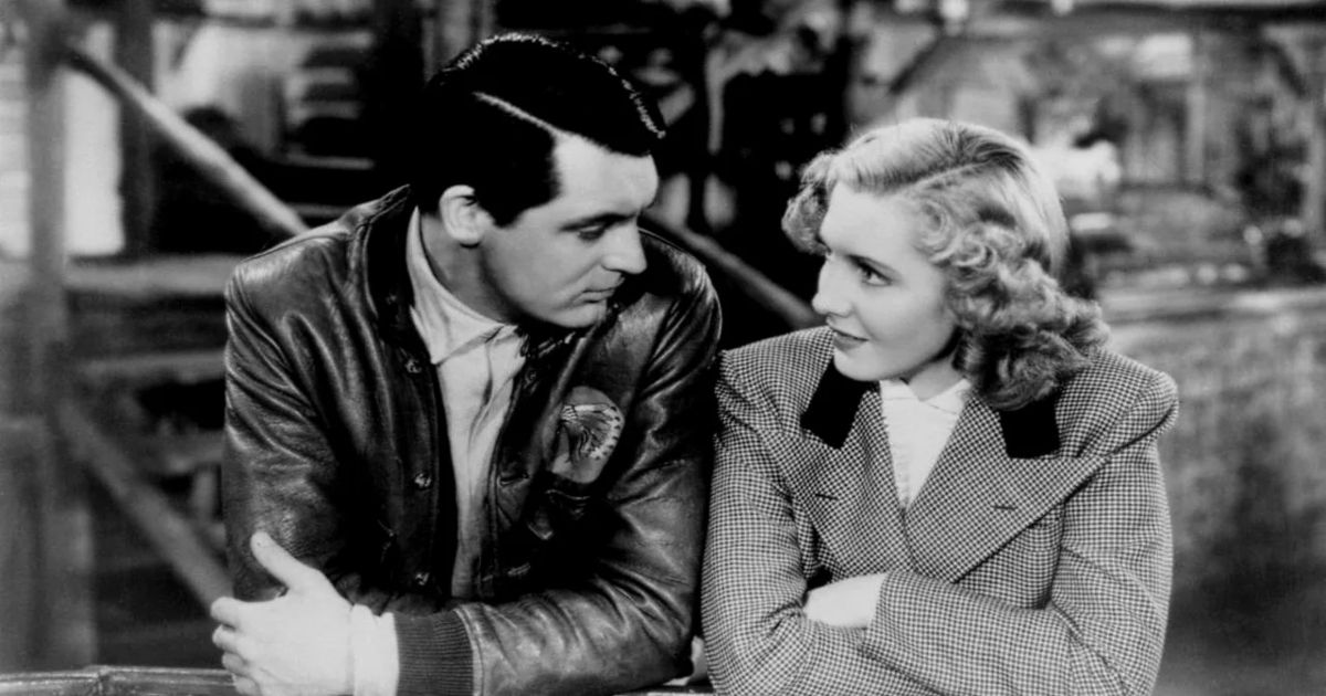 Cary Grant in a leather flight jacket leaning against a railing with a blonde in Only Angels Have Wings