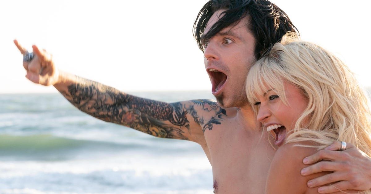 Man with heavily tattooed arm raises it while holding woman; they stand in front of the ocean.
