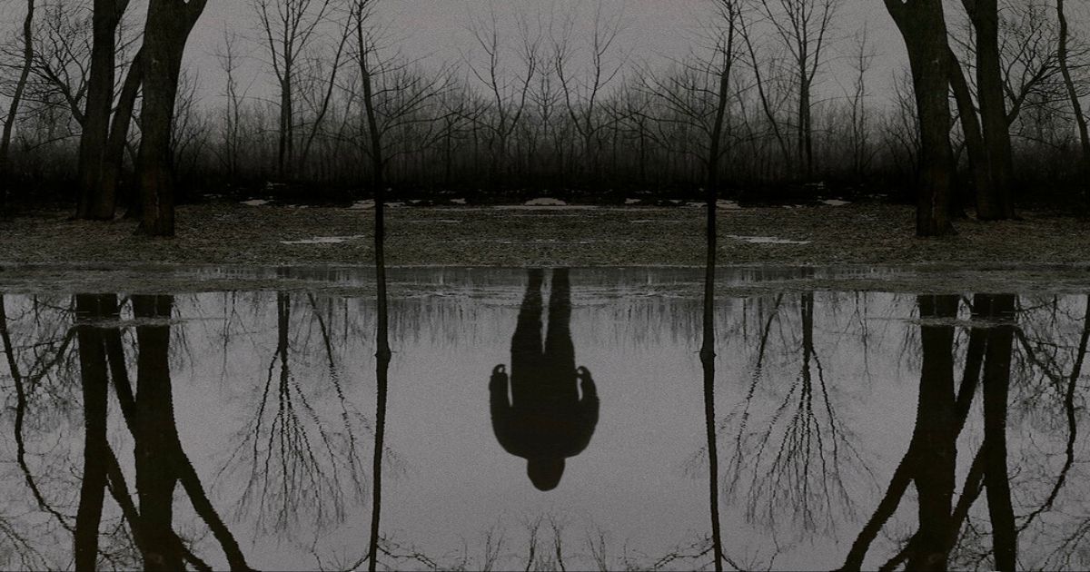 A man stands by a lake with his reflection in it in The Outsider