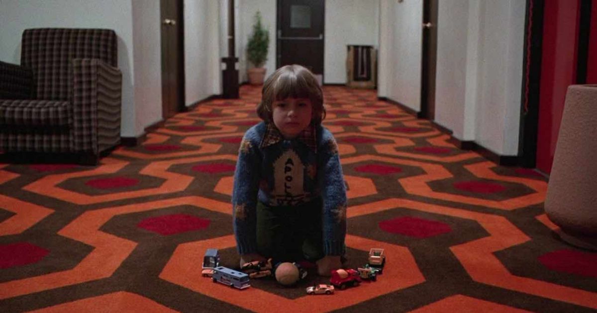 Danny sits in the patterned carpet hallway of the Overlook Hotel in The Shining 
