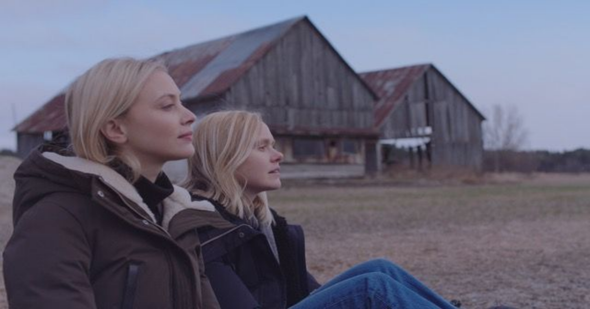Sarah Gadon and Alison Pill sitting on the grass by a barn in All My Puny Sorrows