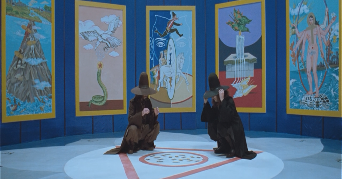 Strange figures sit in a blue room with giant tarot card paintings in The Holy Mountain