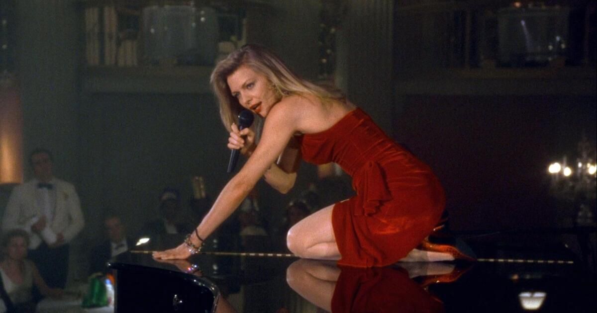 Woman sits atop piano in red dress and heels.