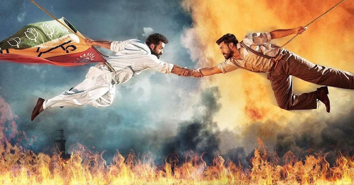 The two main characters of RRR hold each other, wrist-to-wrist, over a river on fire.