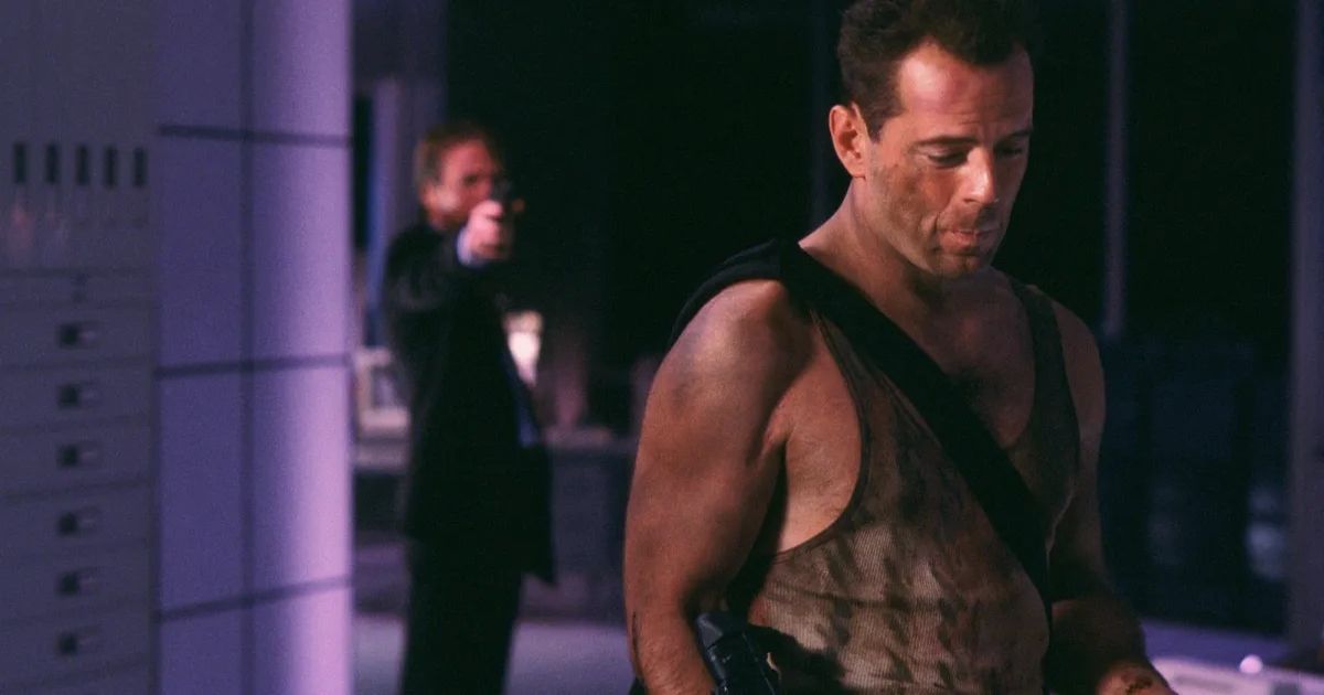Bruce Willis with his back turned to a man pointing a gun at him in Die Hard.