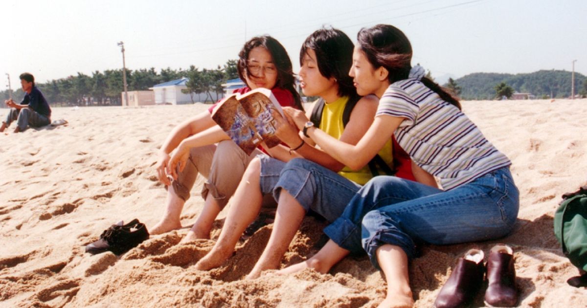 Three girls sit on a beach with a book.