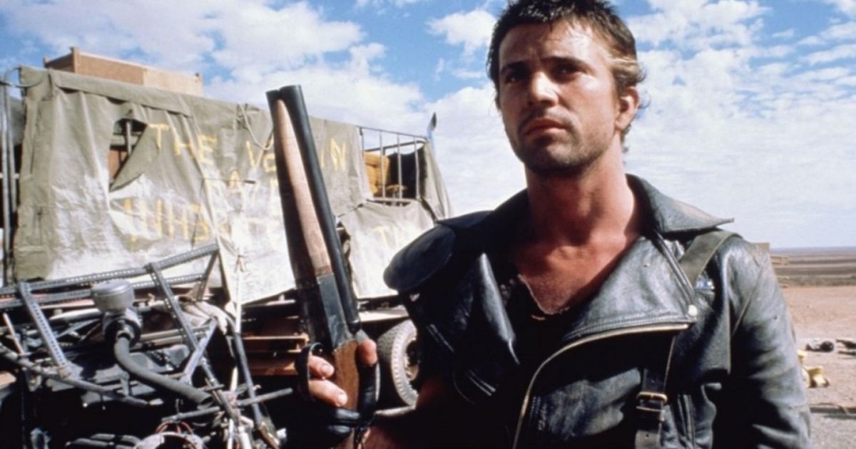 Mad Max is wearing his leather bomber jacket and holding a gun. 