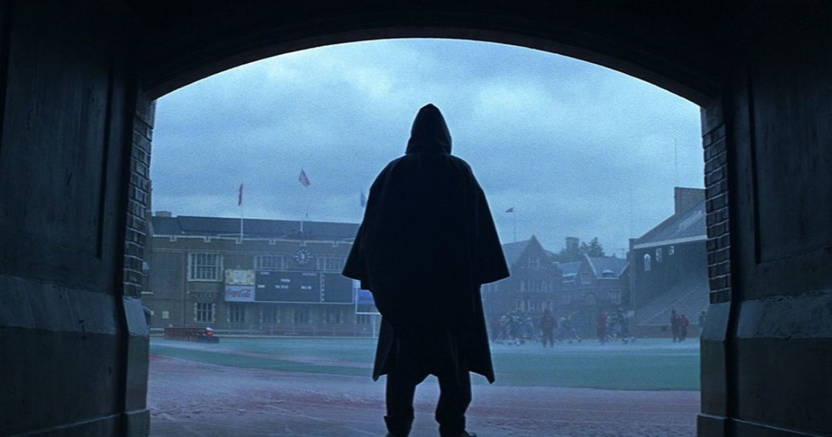 A hooded figure stands at the team entrance of a football field, watching a game in the rain.