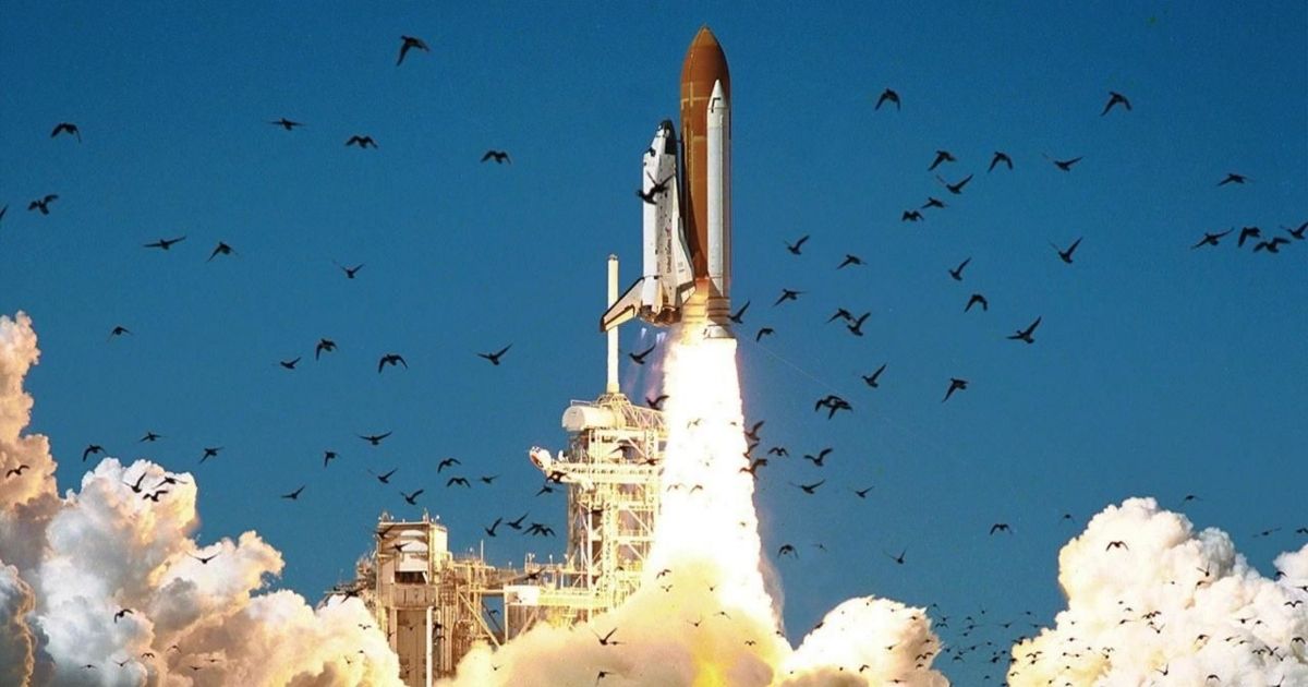 A rocket being shot into space, the smoke billowing below and scaring off a bunch of birds