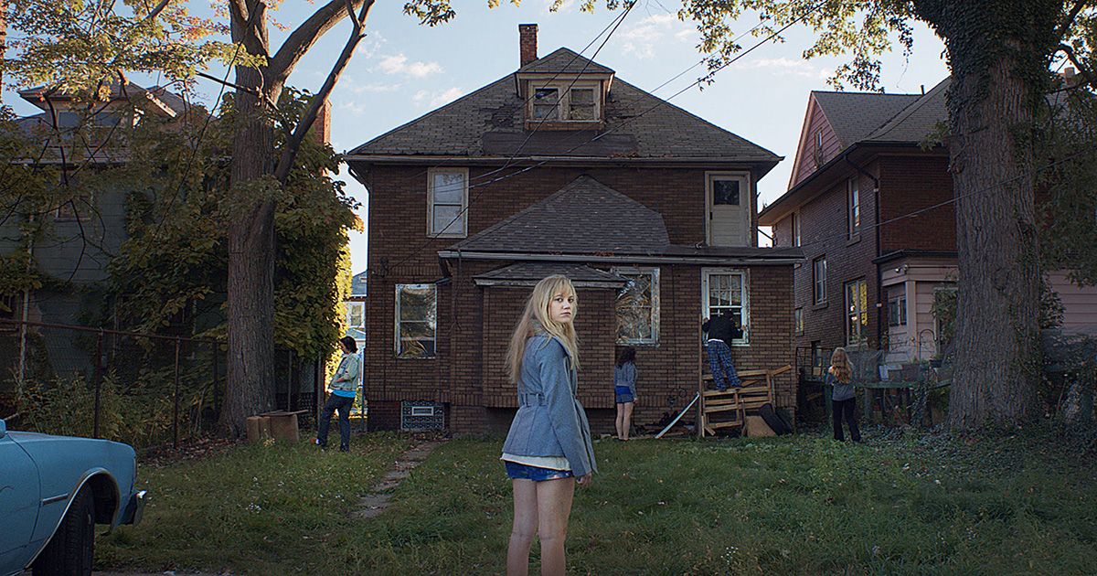 Maika Monroe, as Jay Height, turns around and looks toward the camera as she stands in front of a house with different characters in the distance in It Follows.