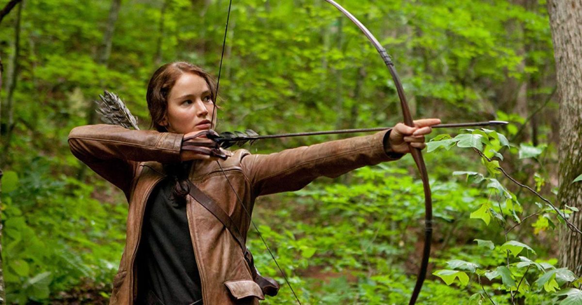 Katniss dans une veste marron tirant son arc et ses flèches dans la forêt verte