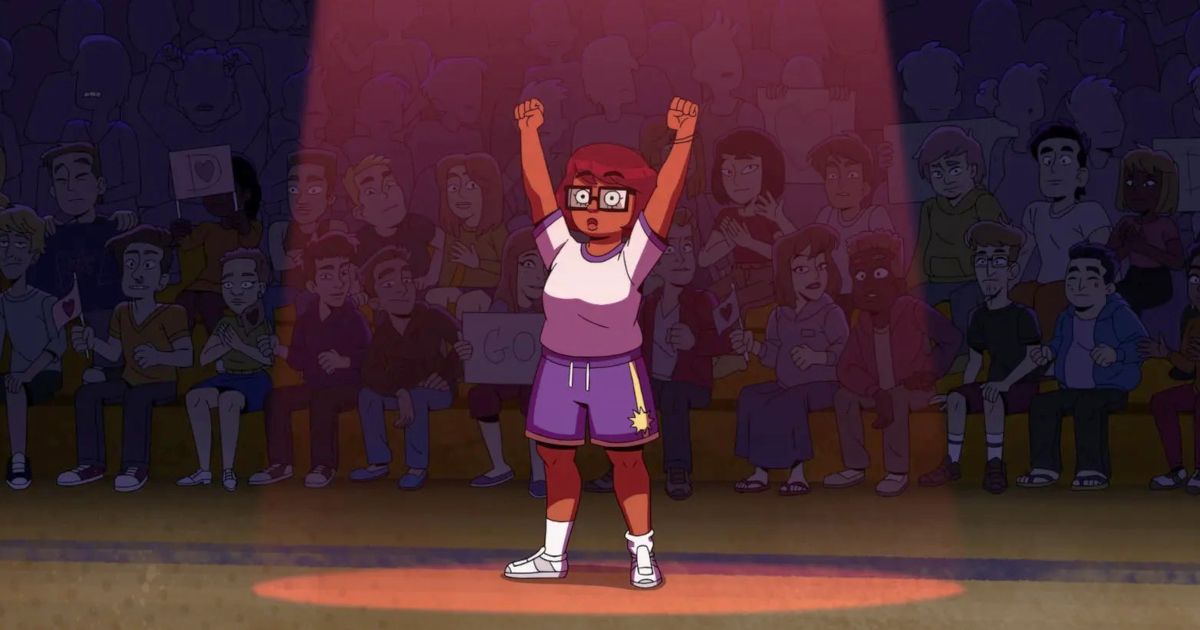 Velma em pé no meio de um ginásio em uma função escolar com os braços para cima.