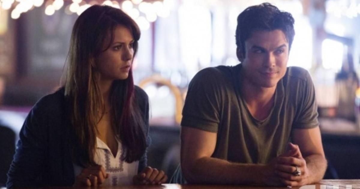 Nina Dobrev and Ian Somerhalder In The Vampire DIaries. Nina as "Elena" leans against a bench next to Ian as "Damon" both leaning and listening to an unseen orator. 