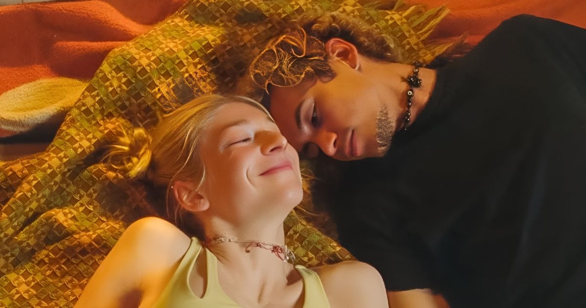 A blonde woman laying down next to a man in a black shirt and blone curls. 