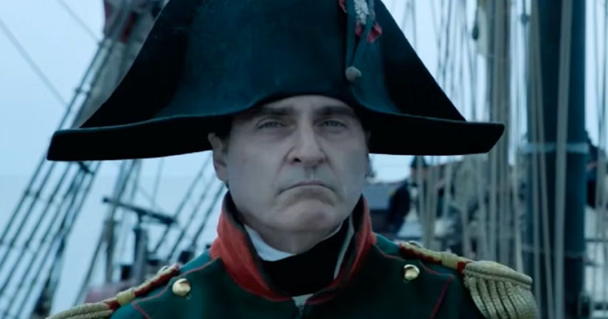 Joaquin Phoenix as Napoleon, wearing a military uniform and large hat while standing ona ship, looking toward the camera with a blank face in Napoleon.