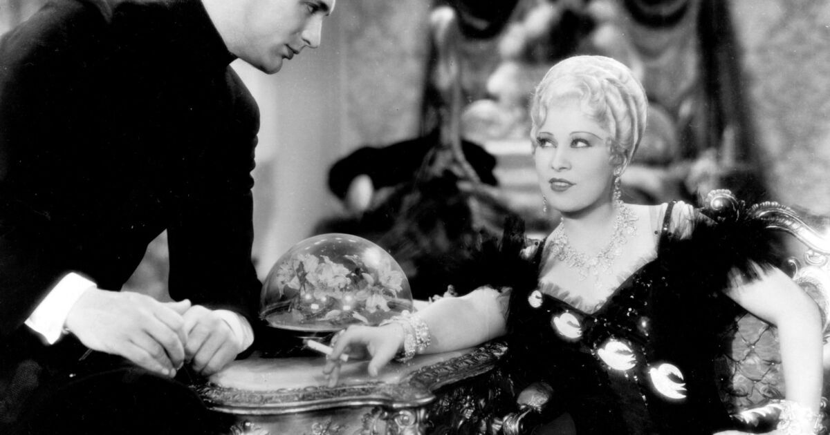 mae west sitting at a table, being approached by a man