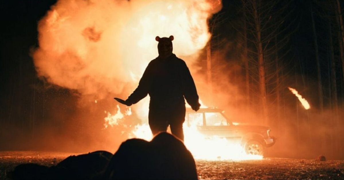 Winnie the Pooh walks with a large knife in his hands towards a victim while a car is burning in the background in Winnie the Pooh: Blood and Honey.