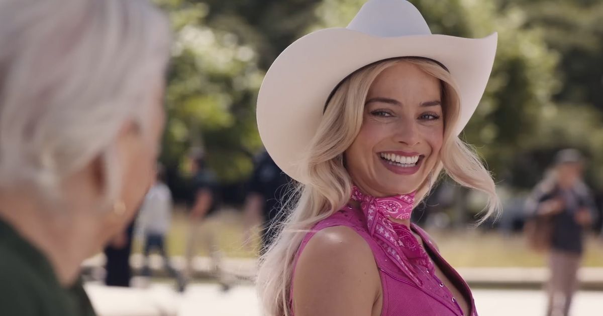 Margot Robbie as Barbie, wearing a cowboy hat, bandana around her neck, and pink outfit, talking to an older woman on a bench in the real world in Barbie.