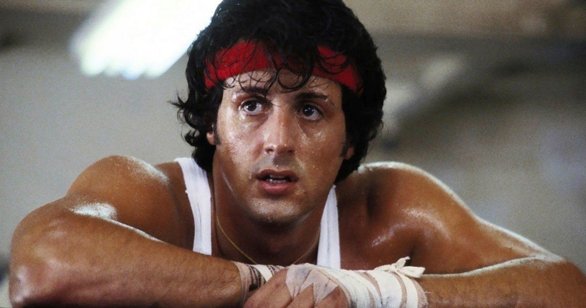 Sylvester Stallone wearing a white tank top and red head band, with his hands taped up in Rocky Balboa
