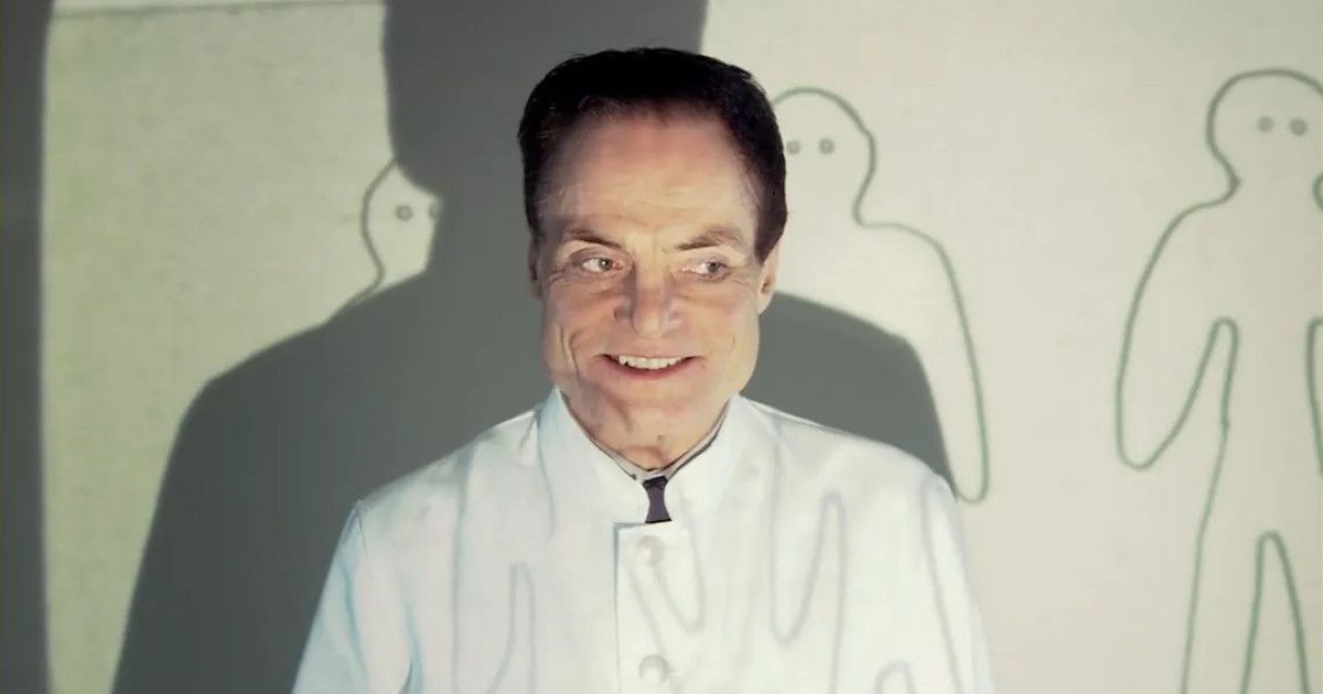 Dieter Laser in a white lab coat giving a presentation in front of a projector with people drawn behind him in The Human Centipede: First Sequence