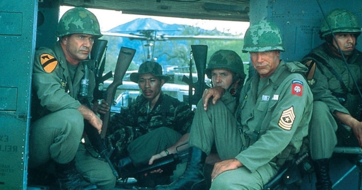 Mel Gibson as Lt. Col. Hal Moore, Sam Elliot as Sgt. Maj. Basil Plumley, with other soldiers in the background, as they all hold guns in their green uniforms and wait on the side of a helicopter in the Vietnam war in We Were Soldiers.