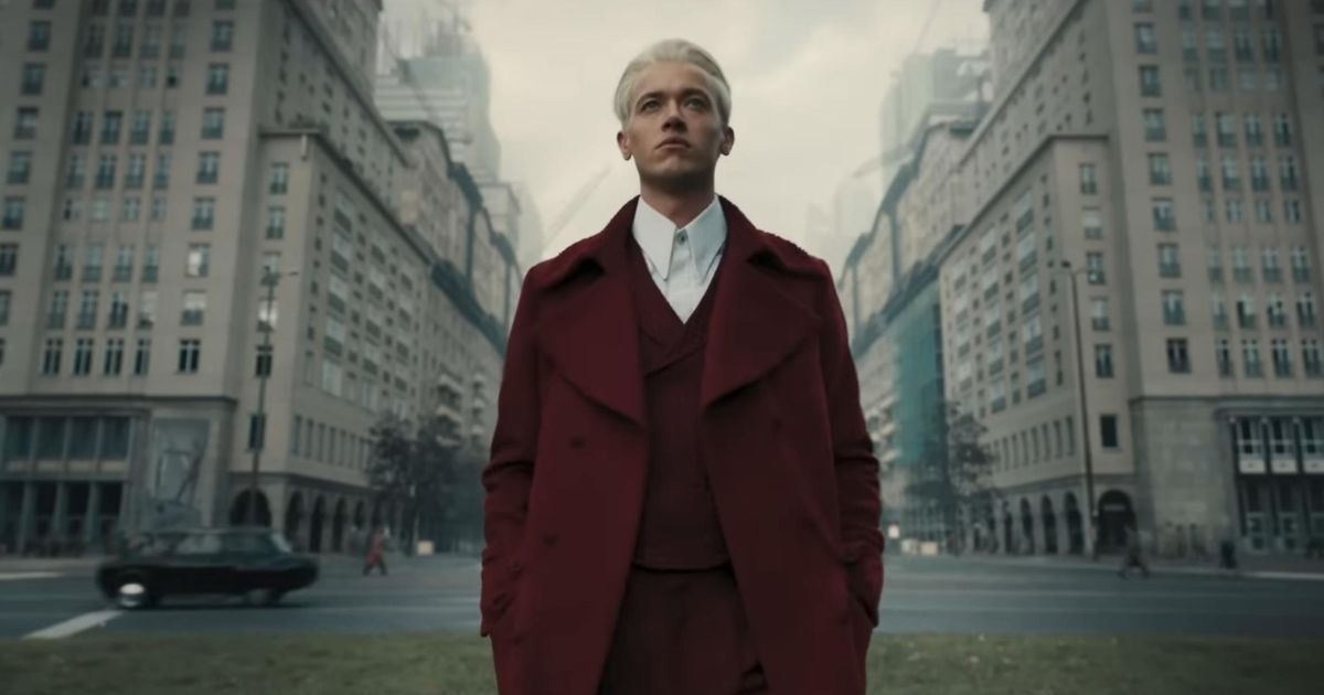 Tom Blyth as Coriolanus Snow standing in the Capitol in a red coat and shirt with a white collared shirt in The Hunger Games: The Ballad of Songbirds and Snakes