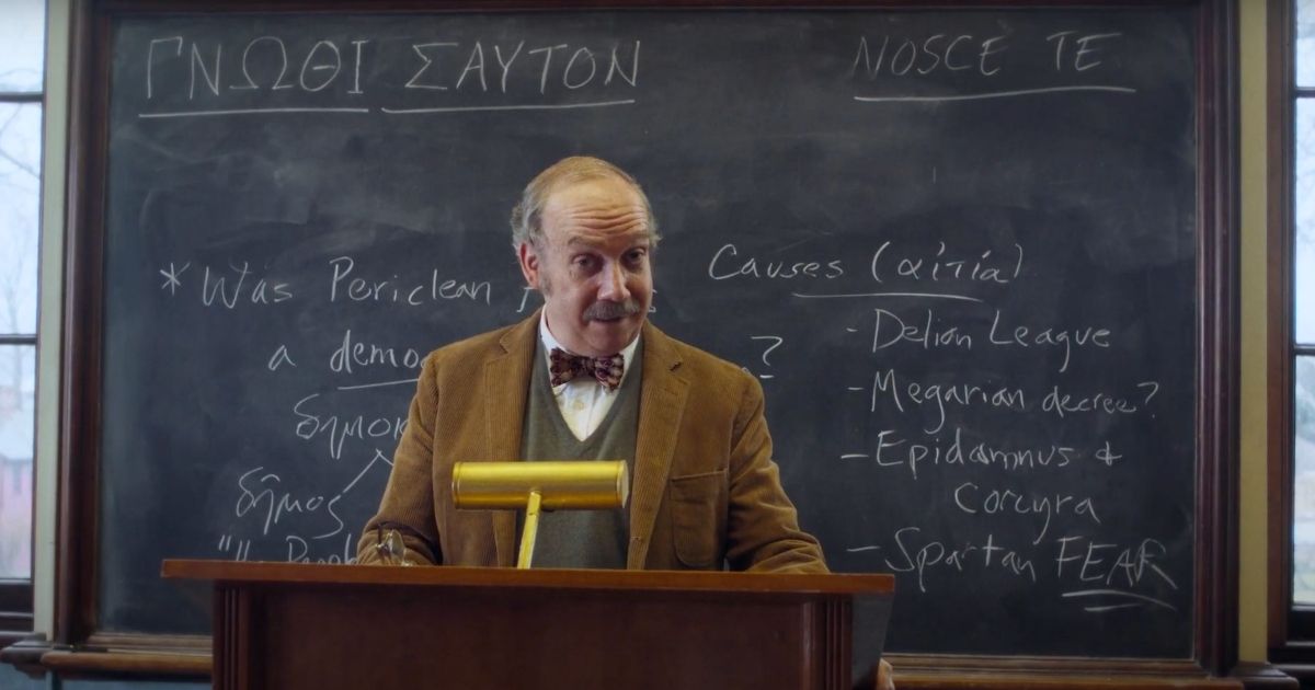 Paul Giamatti in a tan suit jacket, sweater, and shirt wearing a bow tie standing in front of a blackboard with chalk writing on it in a college class in The Holdovers