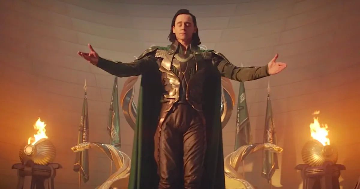 Tom Hiddleston in Loki wearing his green, brown, and black armor as fires burn in his throne room.