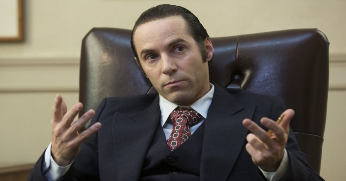 Alessandro Nivola wearing a suit sitting in a large office chair in American Hustle